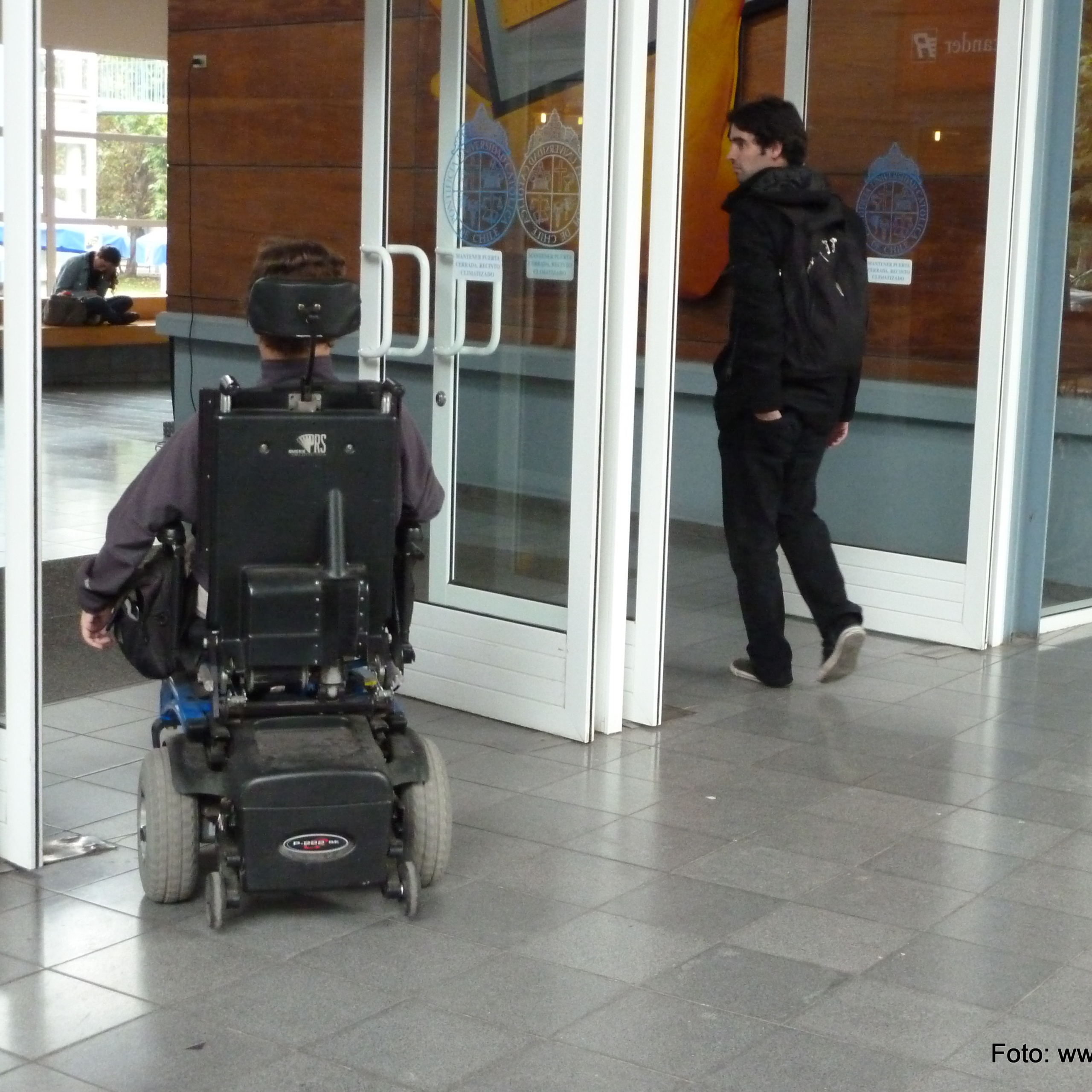 una persona en silla de ruedas y otra caminando cruzan el acceso de una edificación