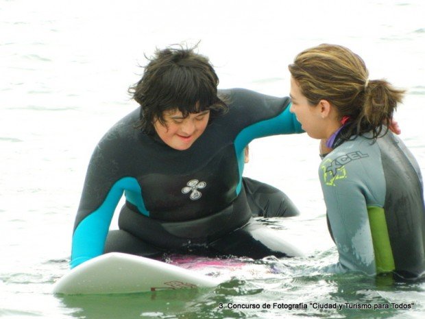 un joven con discapacidad sobre una tabla de surf es acompañado por su instructora