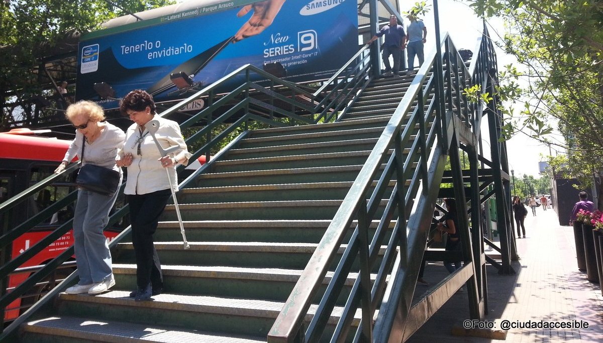 una mujer ayuda a otra mayor a bajar las escaleras de la pasarela peatonal. Una se apoya en el pasamanos y la otra le lleva su bastón