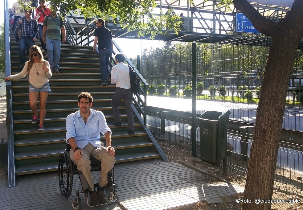 joven en silla de ruedas sentado de espaldas a la escalera de la pasarela peatonal. Varias personas suben y bajan por la escalera