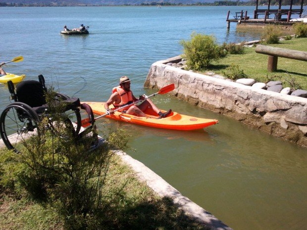 Persona manejando un kayak en una laguna. En primer plano se ve su silla de ruedas esperando en la orilla