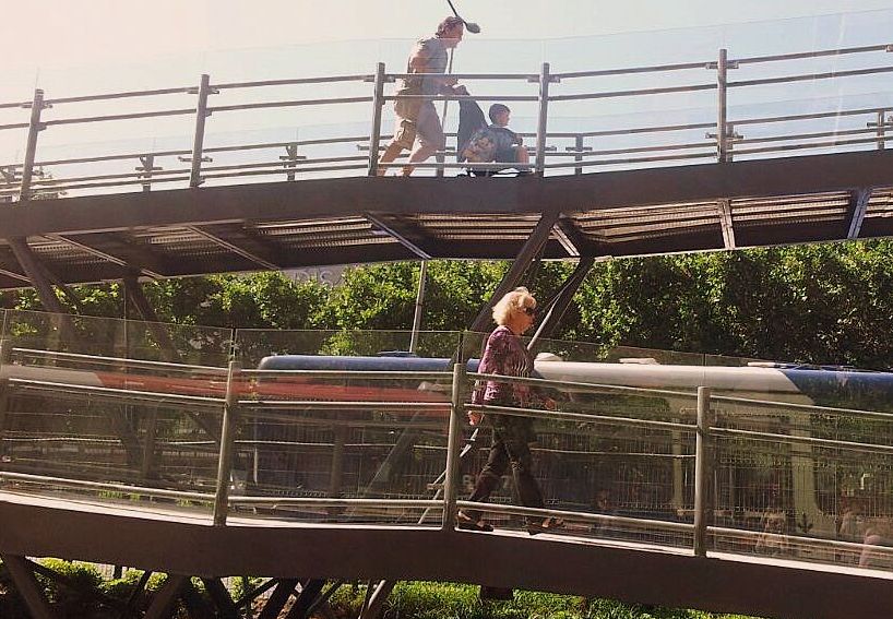 un hombre empuja un coche de niños y en otro nivel camina una mujer mayor sobre la rampa de la pasarela peatonal