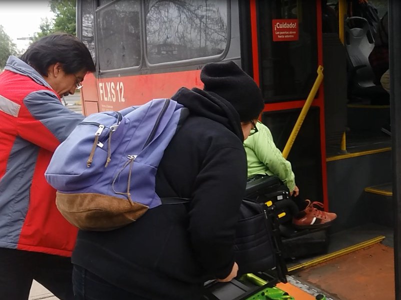 dos personas ayudan a otra en silla de ruedas para acceder al bus