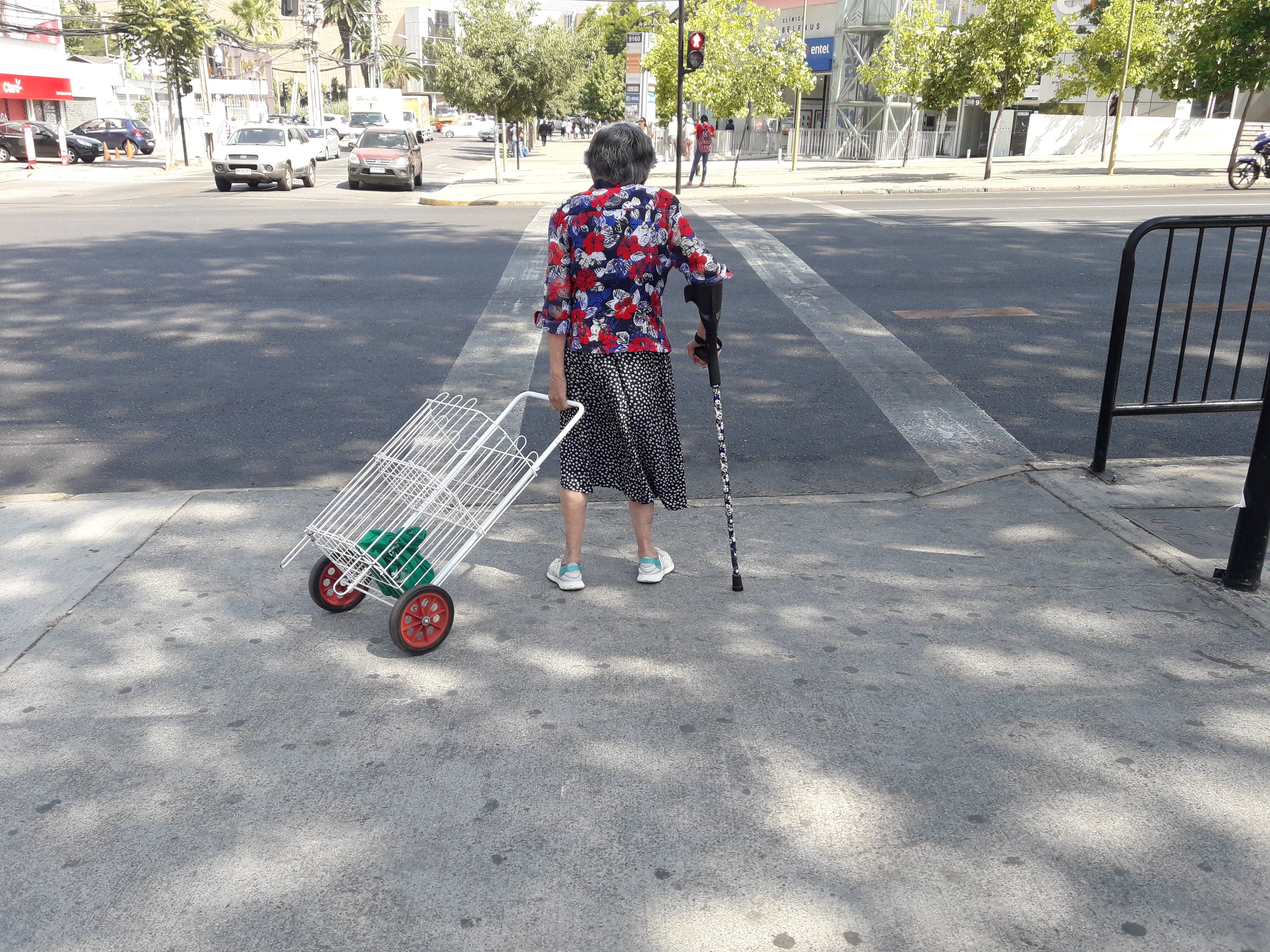 Persona mayor con bastón y un carro de compras enfrentando un cruce peatonal