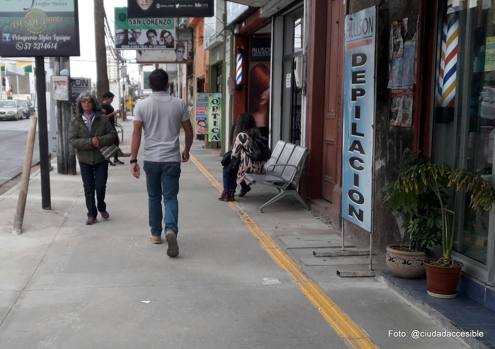 guía podotáctil de avance a lo largo de una vereda donde se ven asientos puestos sobre ésta y carteles