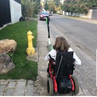 Joven en silla de ruedas por una vereda angosta enfrentando un scooter estacionado en la mitad