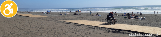 Larga pasarela de madera que recorre la playa de Santo Domingo se interrumpe en un tramo. Una persona en silla de ruedas mira hacia la playa.