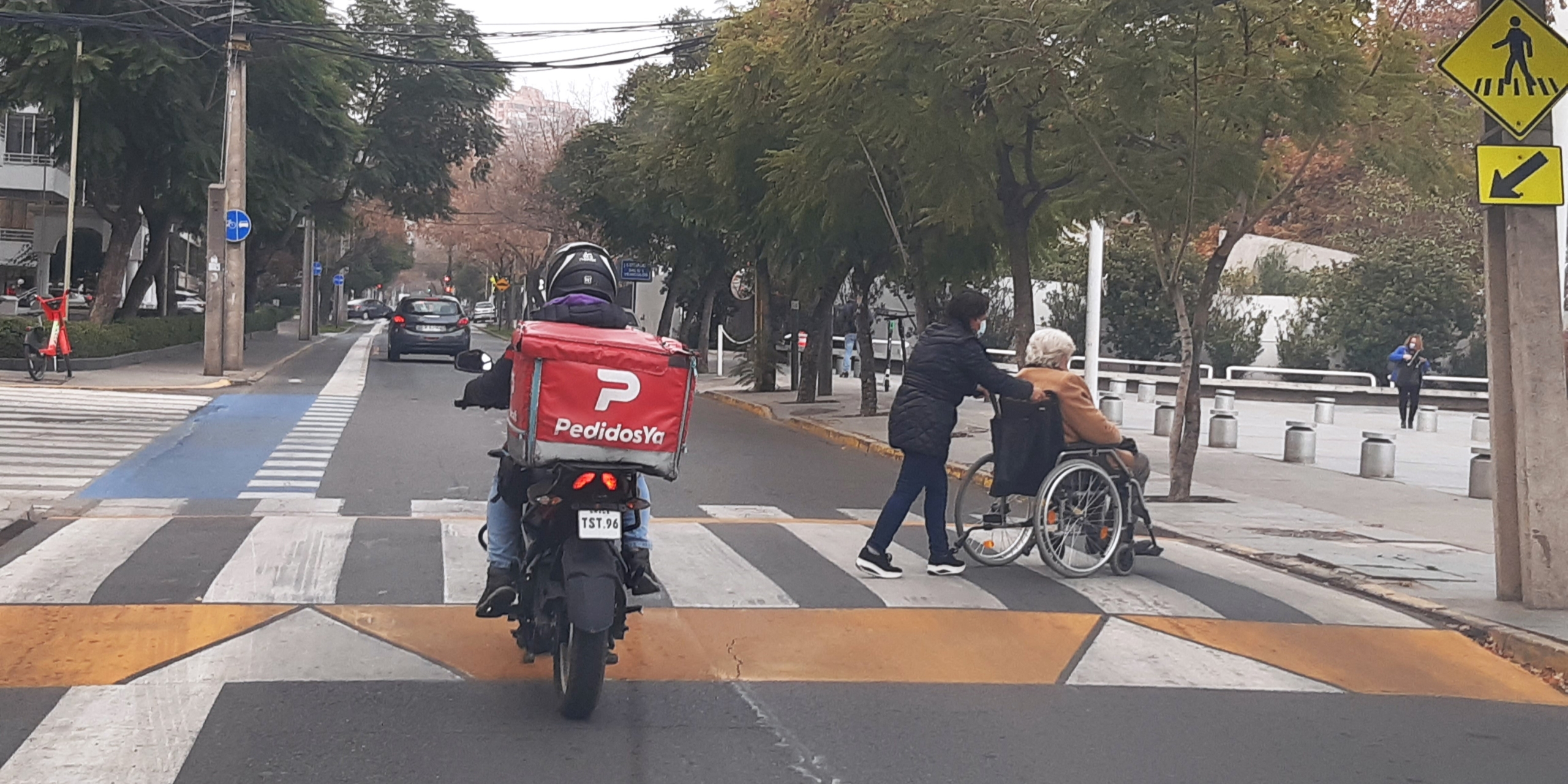 Una persona mayor en silla de ruedas es empujada por su asistente sobre un cruce peatonal a nivel vereda