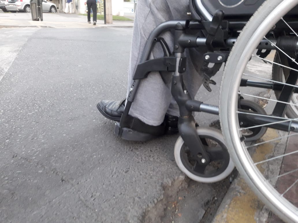 Detalle de las ruedas de una silla de ruedas al enfrentar el mal estado del pavimento en el encuentro de la rampa del rebaje con la calzada.