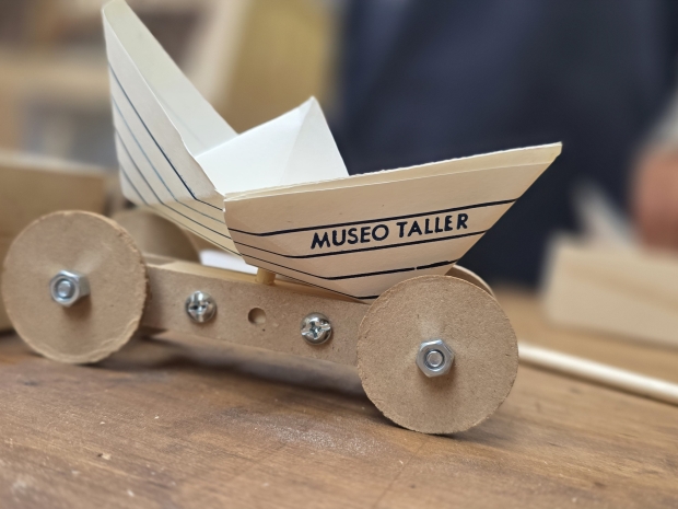 Juguete de madera que consiste en 4 pequeñas ruedas unidas por maderas centrales. Sobre el carrito va un barco de papel